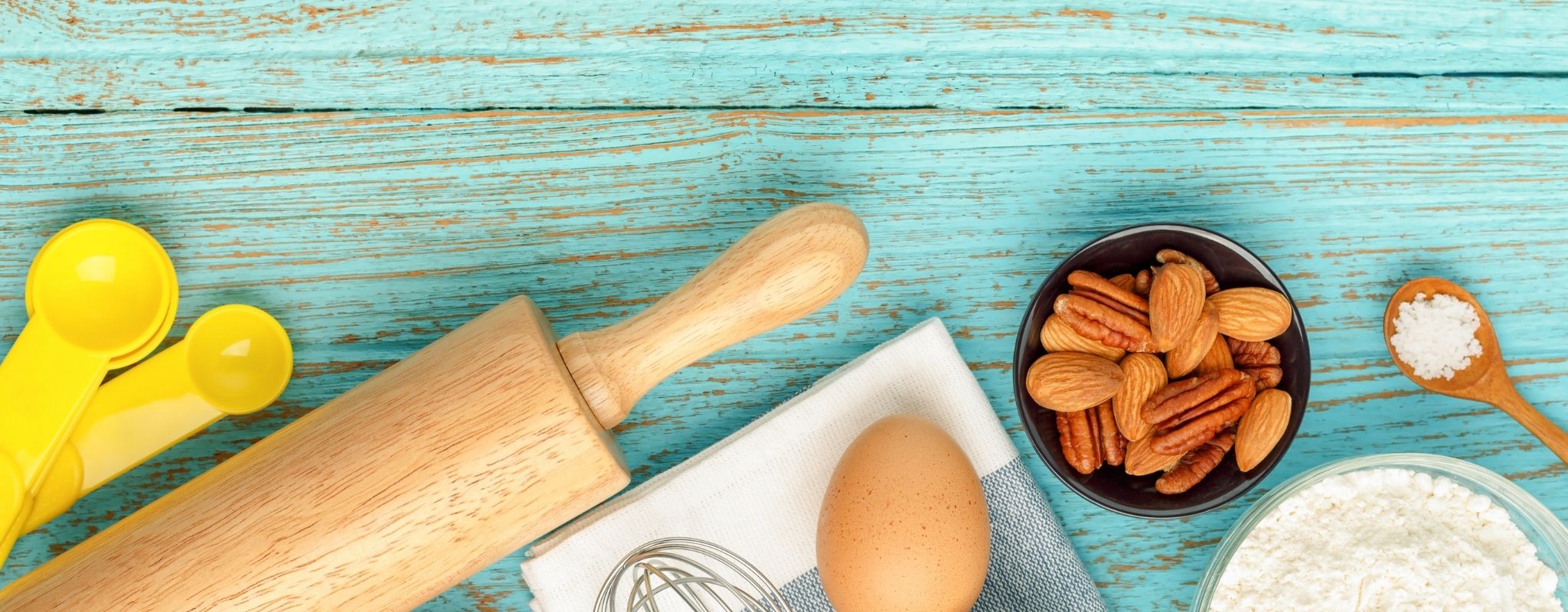 Flat lay of baking ingredients on a wooden table, including flour, butter, eggs, milk, and kitchen tools with a blank recipe notebook.